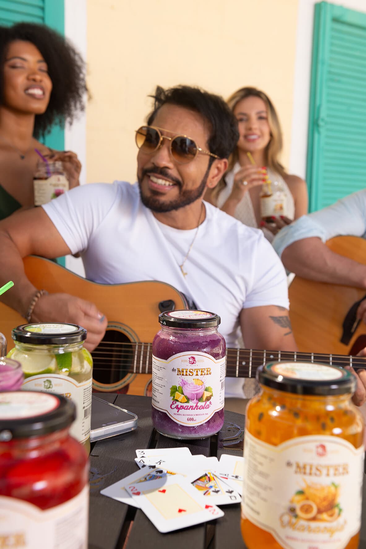 Potes de Mister de espanhola, limão, maracujá e morango na mesa, com amigos e violão ao fundo